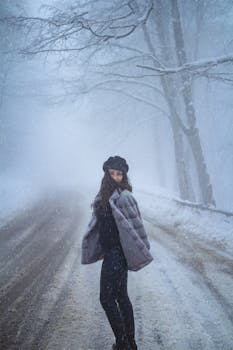Elegant woman in a winter coat and hat standing on a snowy road lined with trees.