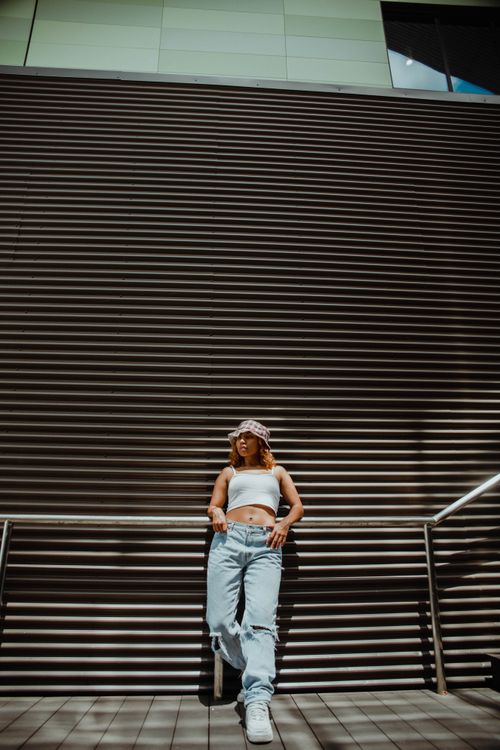 Free From below of young female in casual clothes and hat standing leaning against rail next to textures wall in sunny day Stock Photo