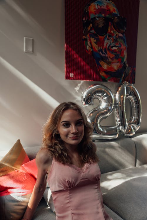 Cheerful young skinny female in pink shirt looking at camera with smile while relaxing on sofa in bright room