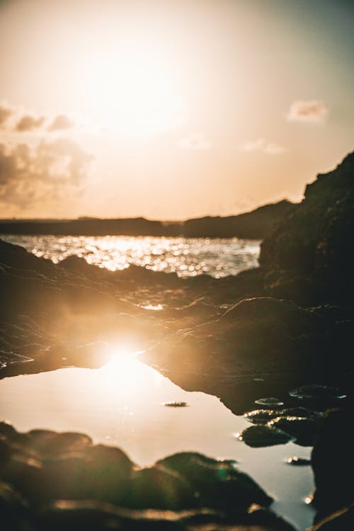 Picturesque view of rippling ocean water near rough coast and puddle with reflection of sundown