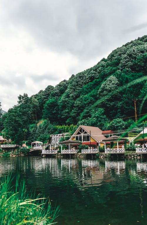 Picturesque landscape of calm river water with houses on coast and lush forest on mountain under cloudy sky