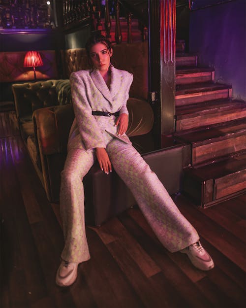 Full body of gorgeous female in elegant suit and sneakers looking at camera while resting on coffee table near sofa and wooden stairway in dark cozy cafe
