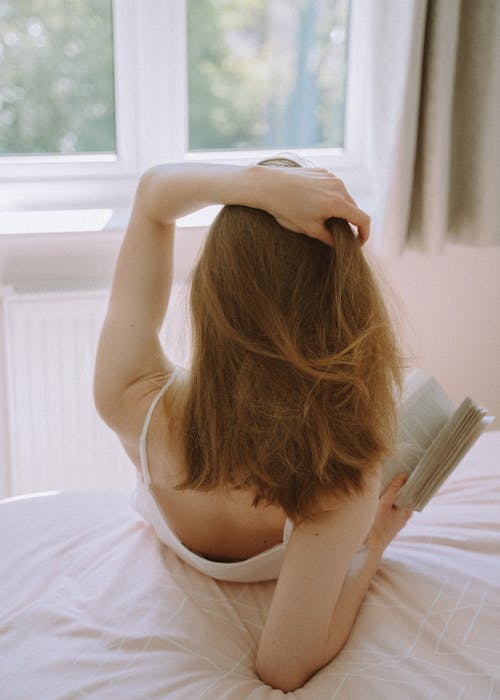 Free  Back View of a Woman Sitting on a Bed while Holding a Book Stock Photo