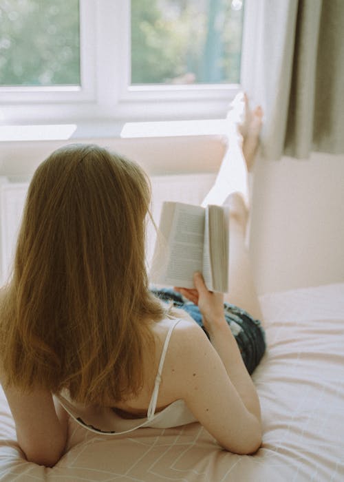 Free Back view of unrecognizable female lying on soft bed and reading interesting book while resting under sunlight Stock Photo