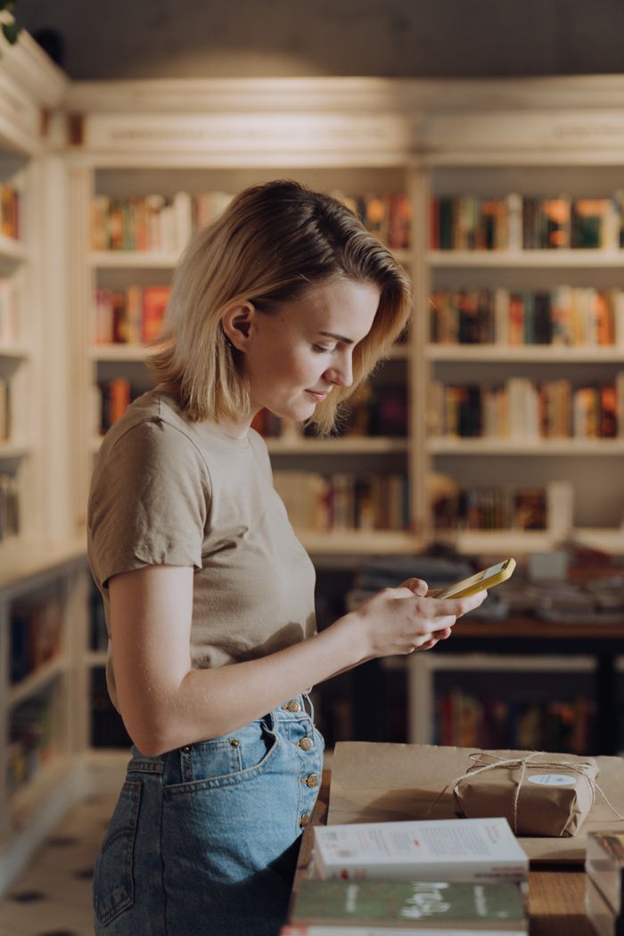 Woman using her phone among library shelves — photo by cottonbro studio on Pexels