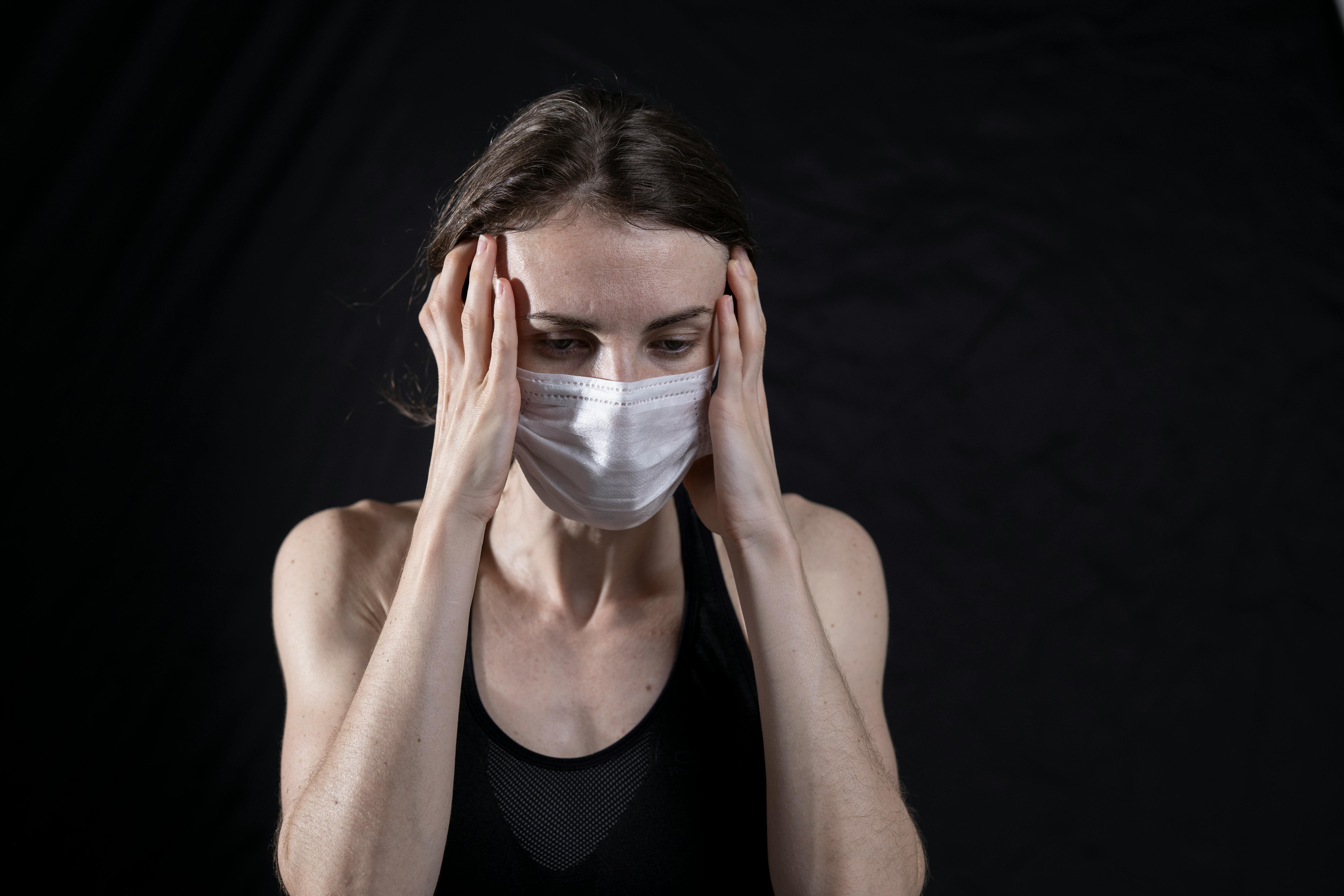 Woman in a face mask holding her temples, conveying stress and anxiety against a black background.