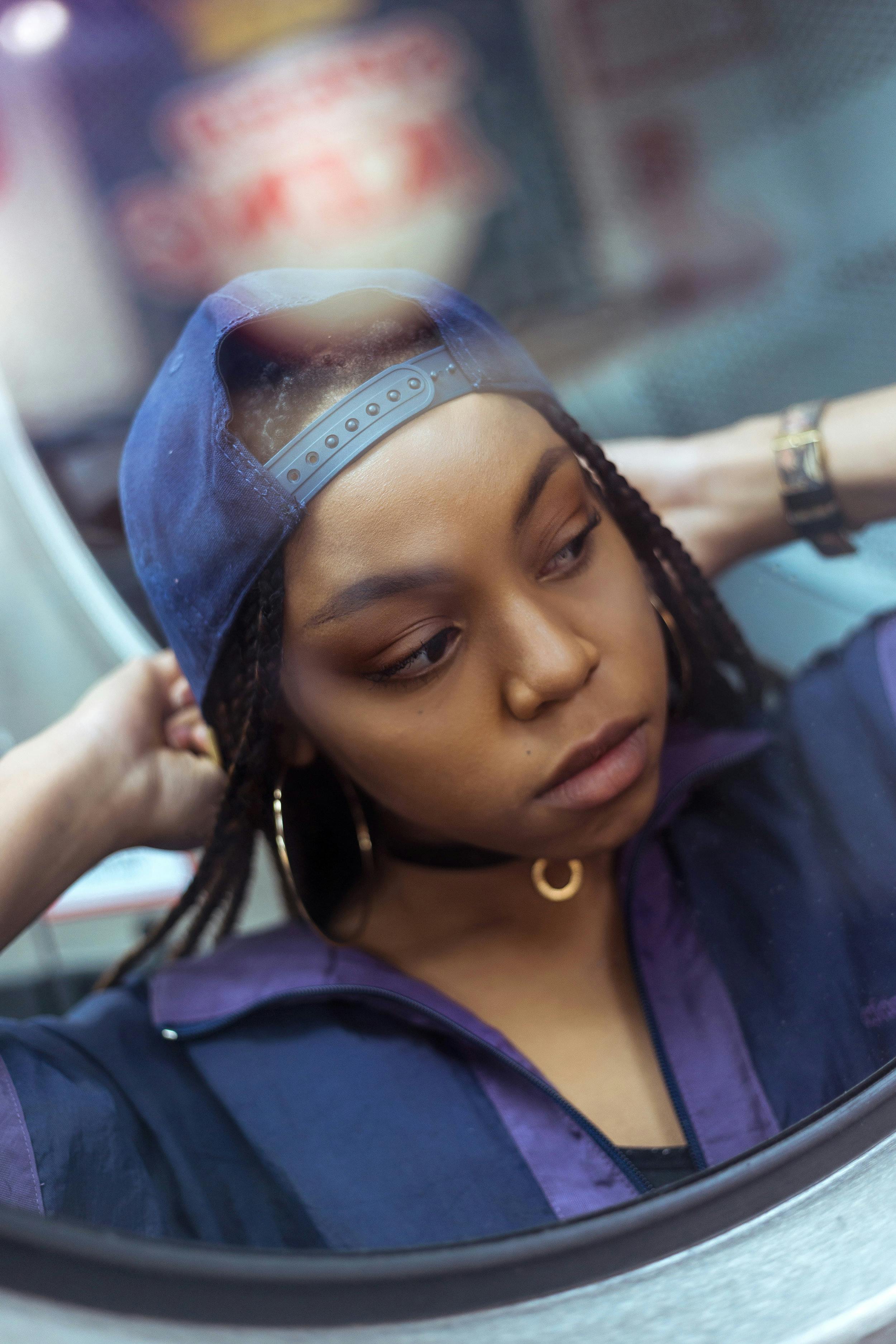 A young woman wearing a cap poses with a retro vibe in an indoor setting, exuding a contemplative mood.
