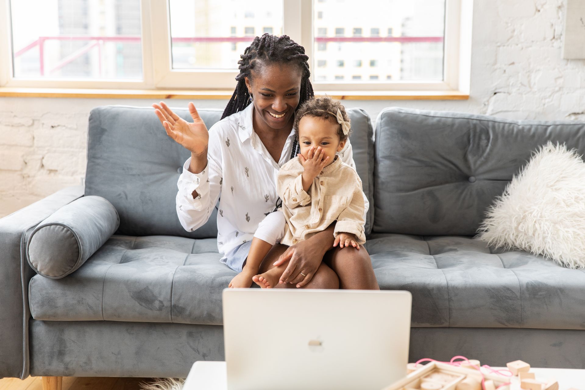 A joyful mother and daughter wave during a video call from their cozy living room couch.