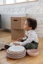 Boy Sitting on Floor by Ceramic Plates and Cardboard Boxes