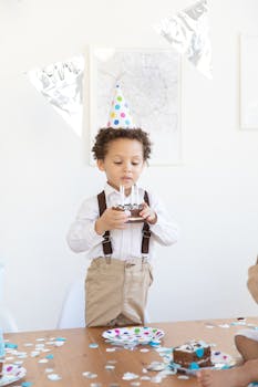 Cute child with party hat holding a birthday cake indoors, celebrating joyfully.