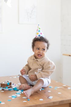 A cute toddler at a birthday party wearing a colorful cap surrounded by cake and confetti indoors.