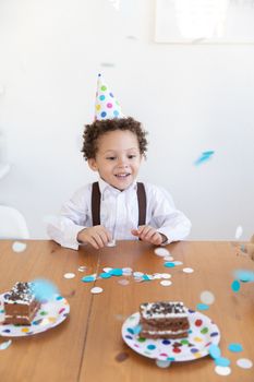Smiling child wearing a party hat surrounded by confetti and cake on a wooden table.