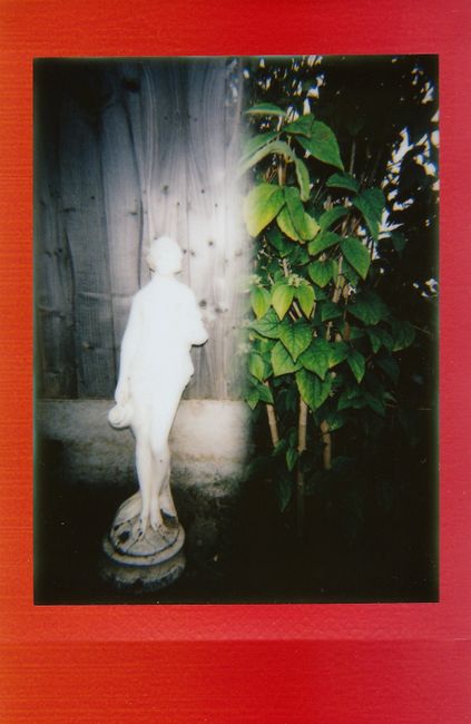 A statue stands near a fence, surrounded by greenery, with a vibrant red frame.
