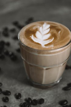 A beautifully crafted latte with leaf art in a glass surrounded by coffee beans on a rustic table.