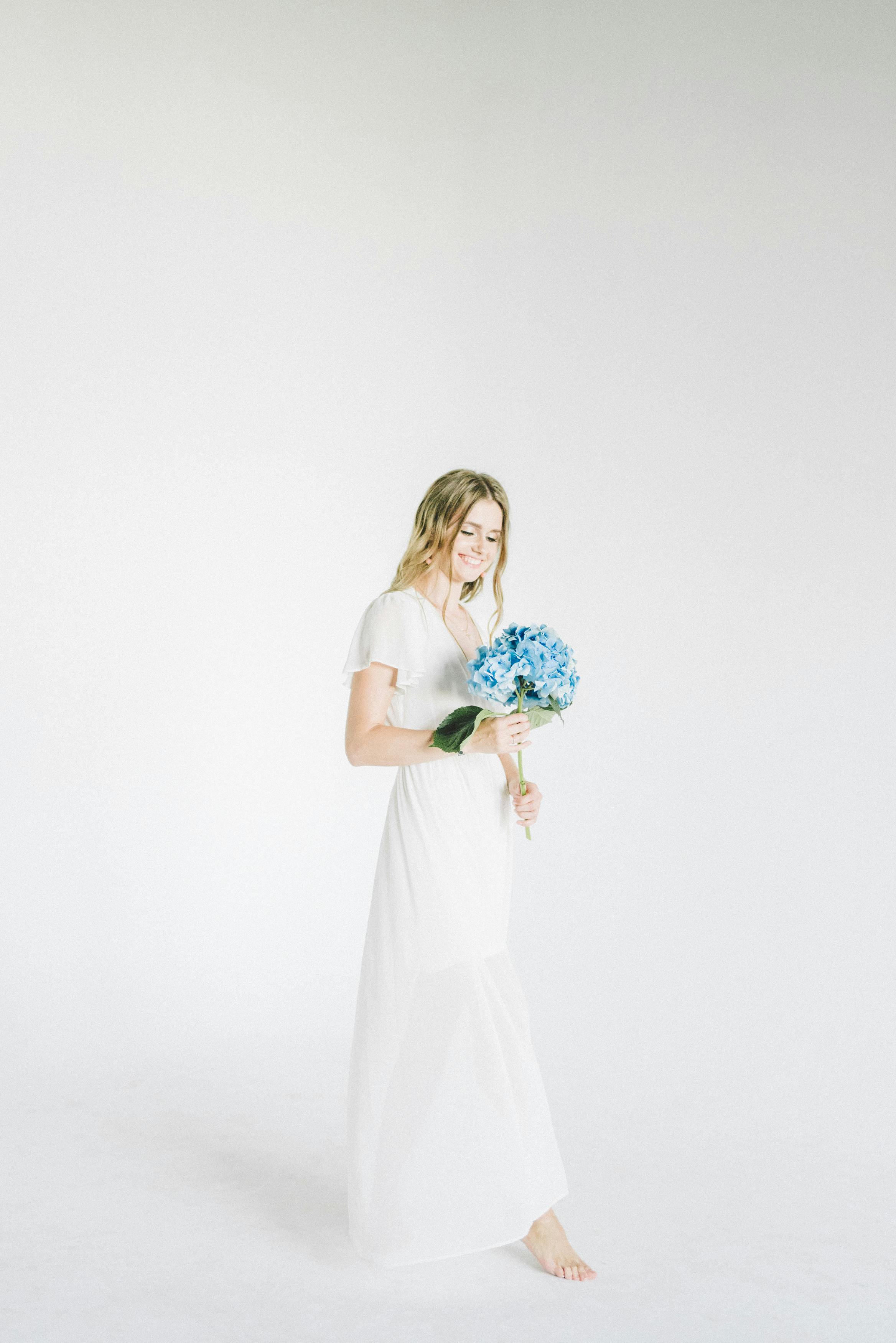 一位身着白色连衣裙的女子手持蓝色绣球花，在明亮环境中宁静地展现肖像