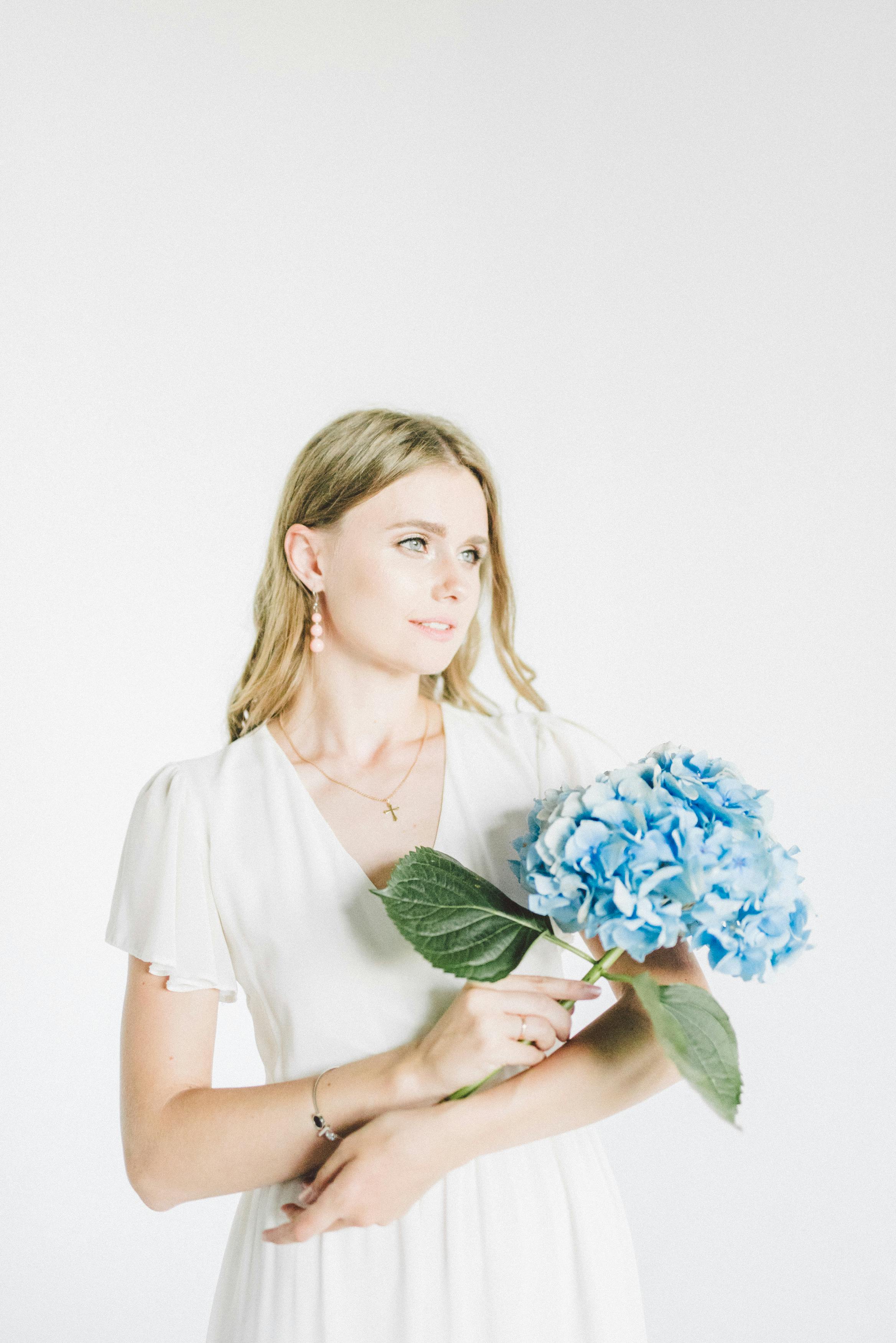 身着白色礼服的女人优雅地站在白色背景前，手持充满活力的蓝色绣球花
