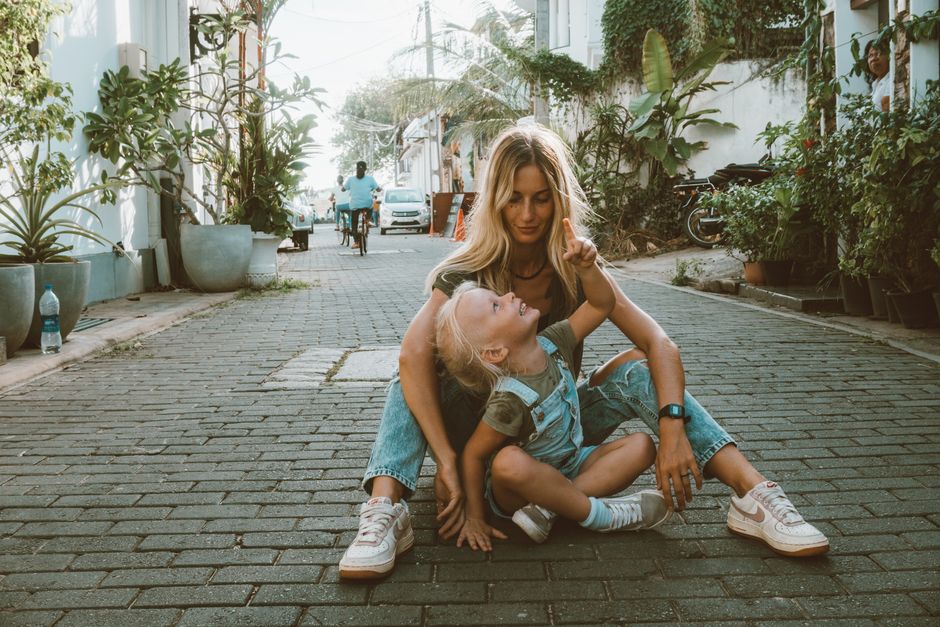 A mother and her daughter enjoying a playful moment on a stone-paved street surrounded by greenery.