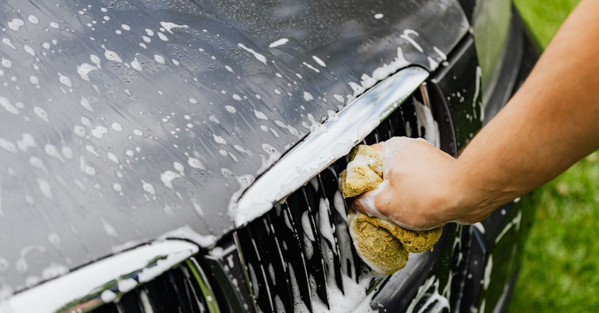 Detailed view of a hand washing a car grille with a soapy sponge outdoors.