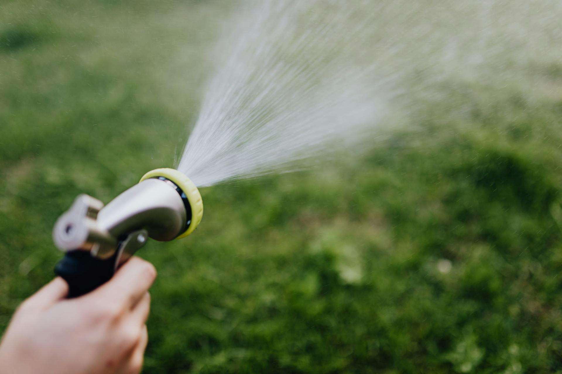 Close-up of a hand using a garden hose sprayer to water the grass outdoors.