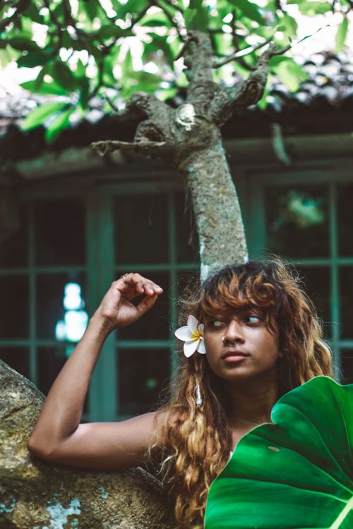 Free A woman with a flower in her hair stands outdoors by a large leaf, showcasing natural beauty. Stock Photo