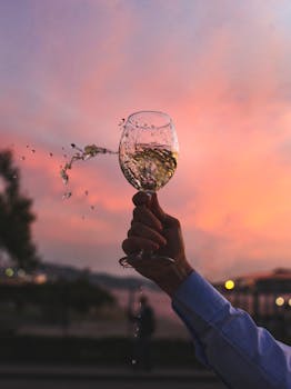 A hand holding a splashing glass of white wine against a stunning sunset backdrop.