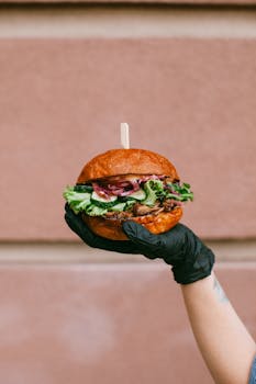 A juicy gourmet burger with fresh toppings held by a gloved hand against a neutral background.