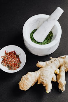 Flat lay of fresh ginger root, herbs, and spices with marble mortar and pestle on dark background.