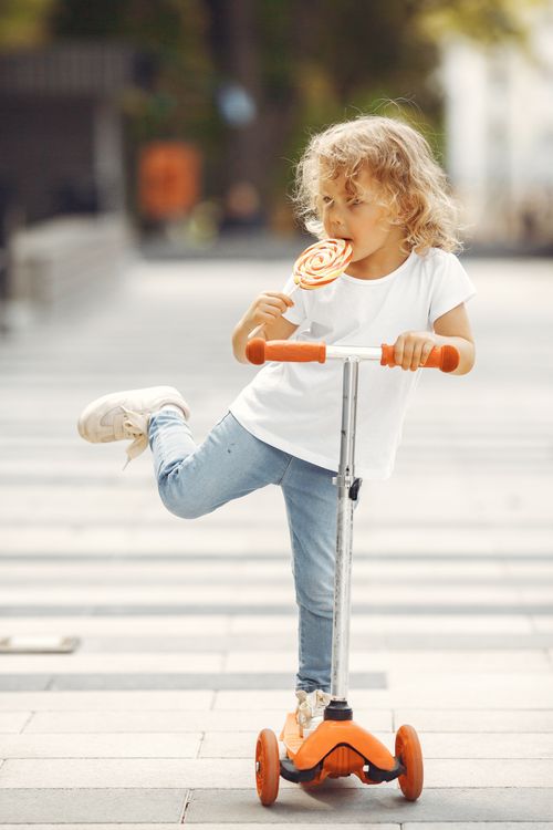 Free A cute child with curly hair enjoys a lollipop while riding a scooter outdoors, exuding innocence and joy. Stock Photo