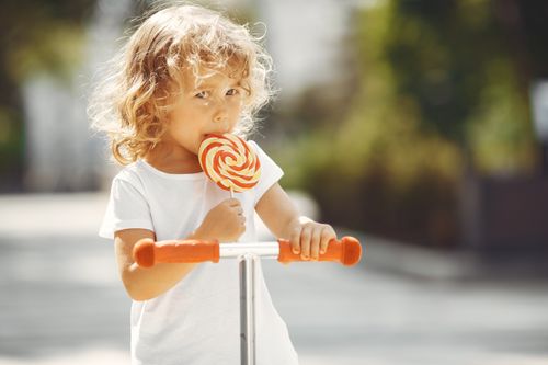 Free Cute young girl enjoying a lollipop while riding a scooter outdoors on a bright sunny day. Stock Photo