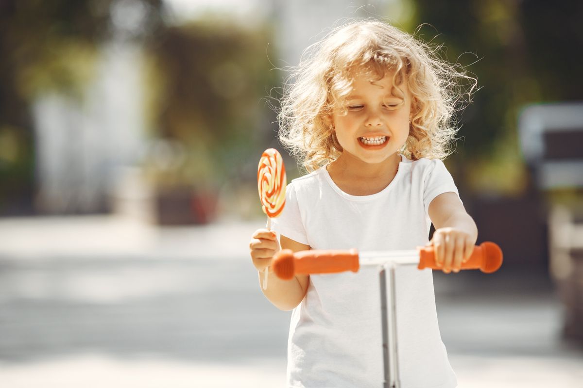 Joyful blonde child rides a scooter while holding a lollipop outdoors on a sunny day.