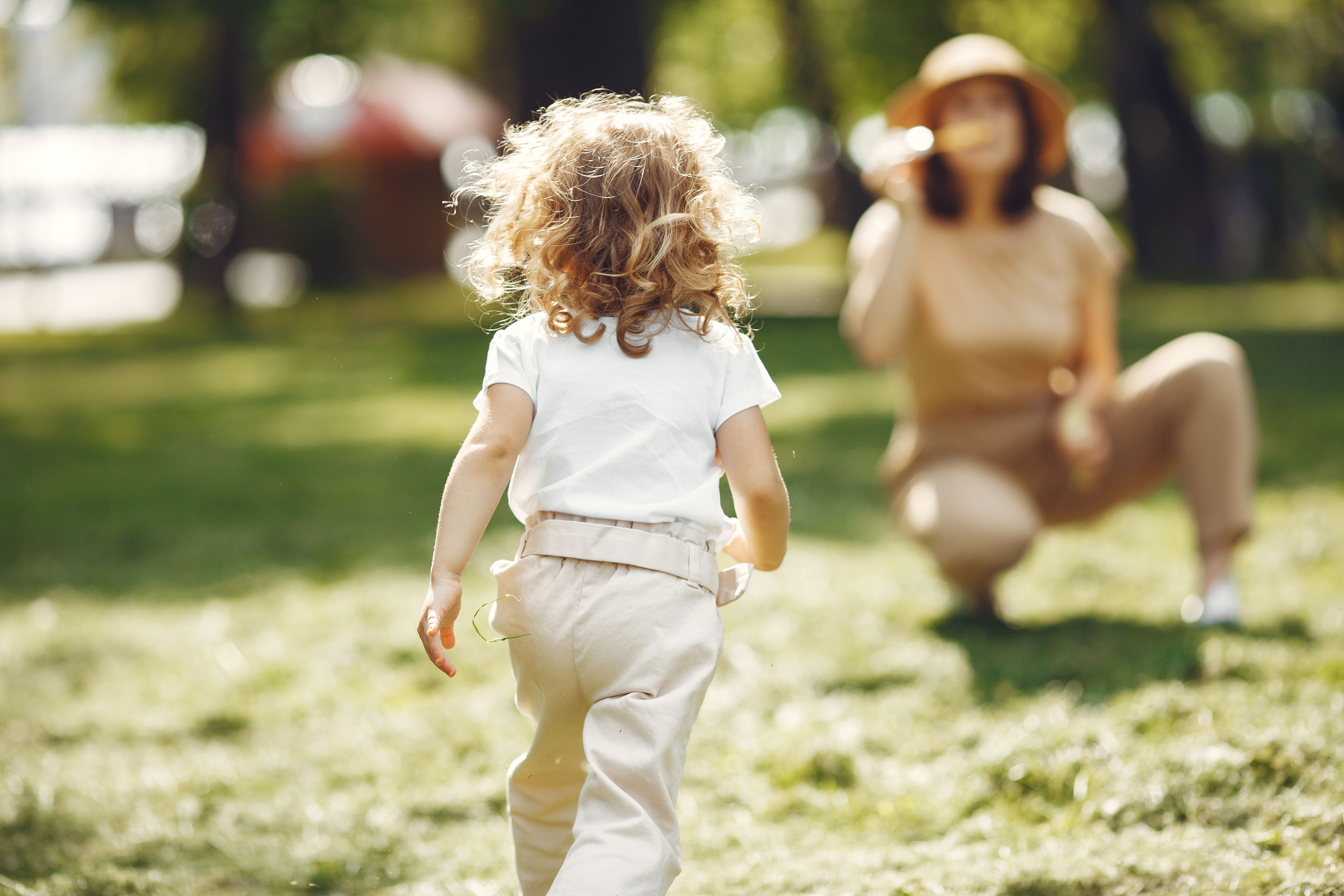 A mother crouches while her child runs towards her in a sunny park.