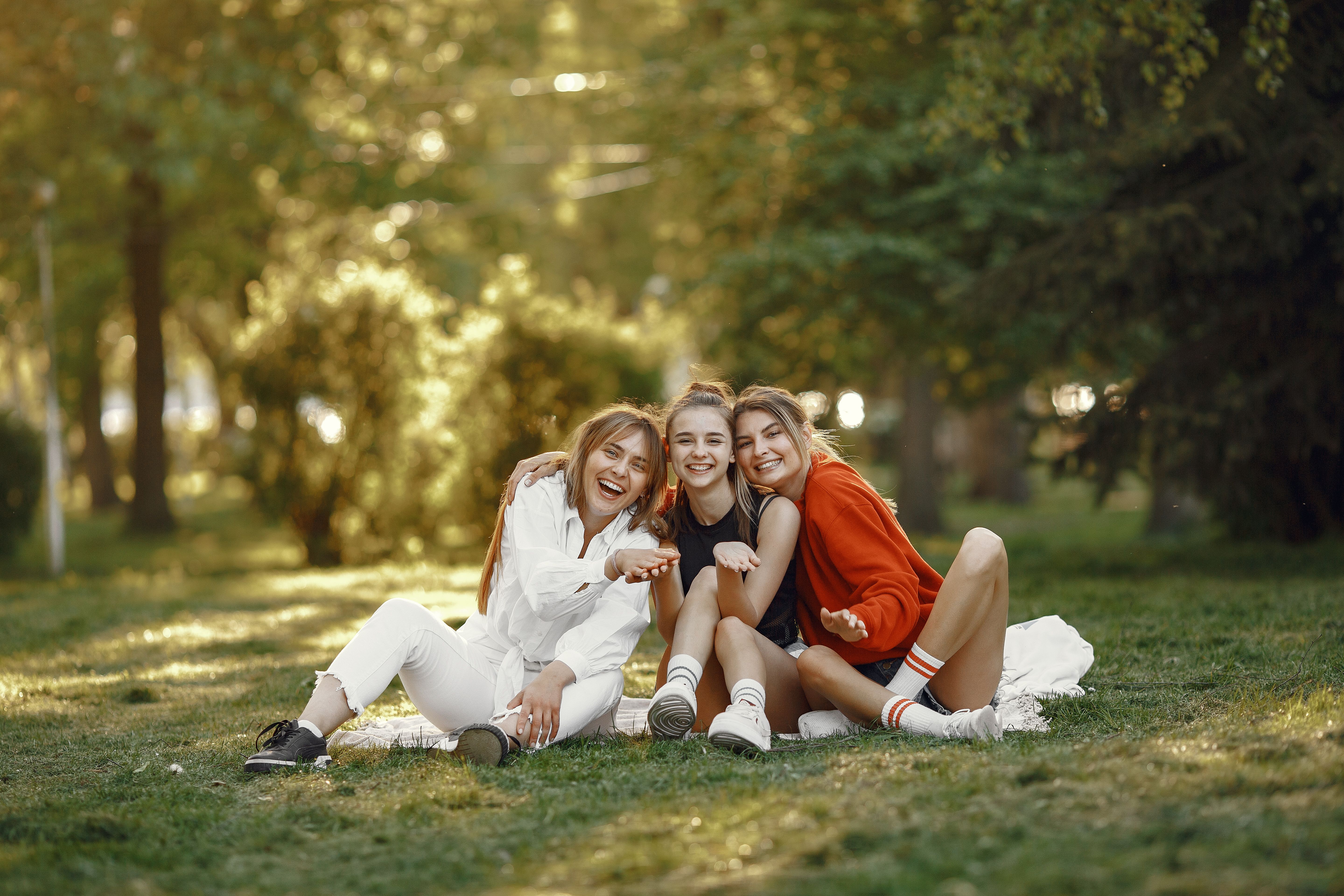 Three happy women sitting together in a sunlit park, laughing and enjoying outdoor leisure time.