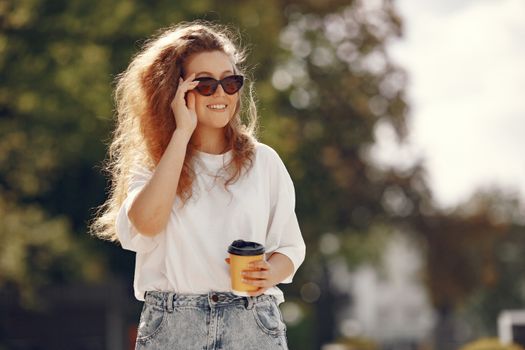 A woman with sunglasses and blonde hair enjoys a coffee in a sunny park.