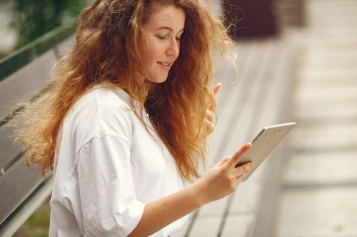 Free Young woman with curly hair reading a tablet while sitting on a bench outside. Stock Photo