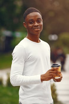 A young man smiling while holding a coffee cup in a park setting.