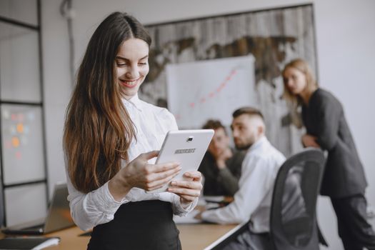 Young woman in a professional office setting browsing a tablet with colleagues in the background.
