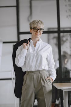 Woman with glasses wearing a white shirt and trousers stands confidently in an office.