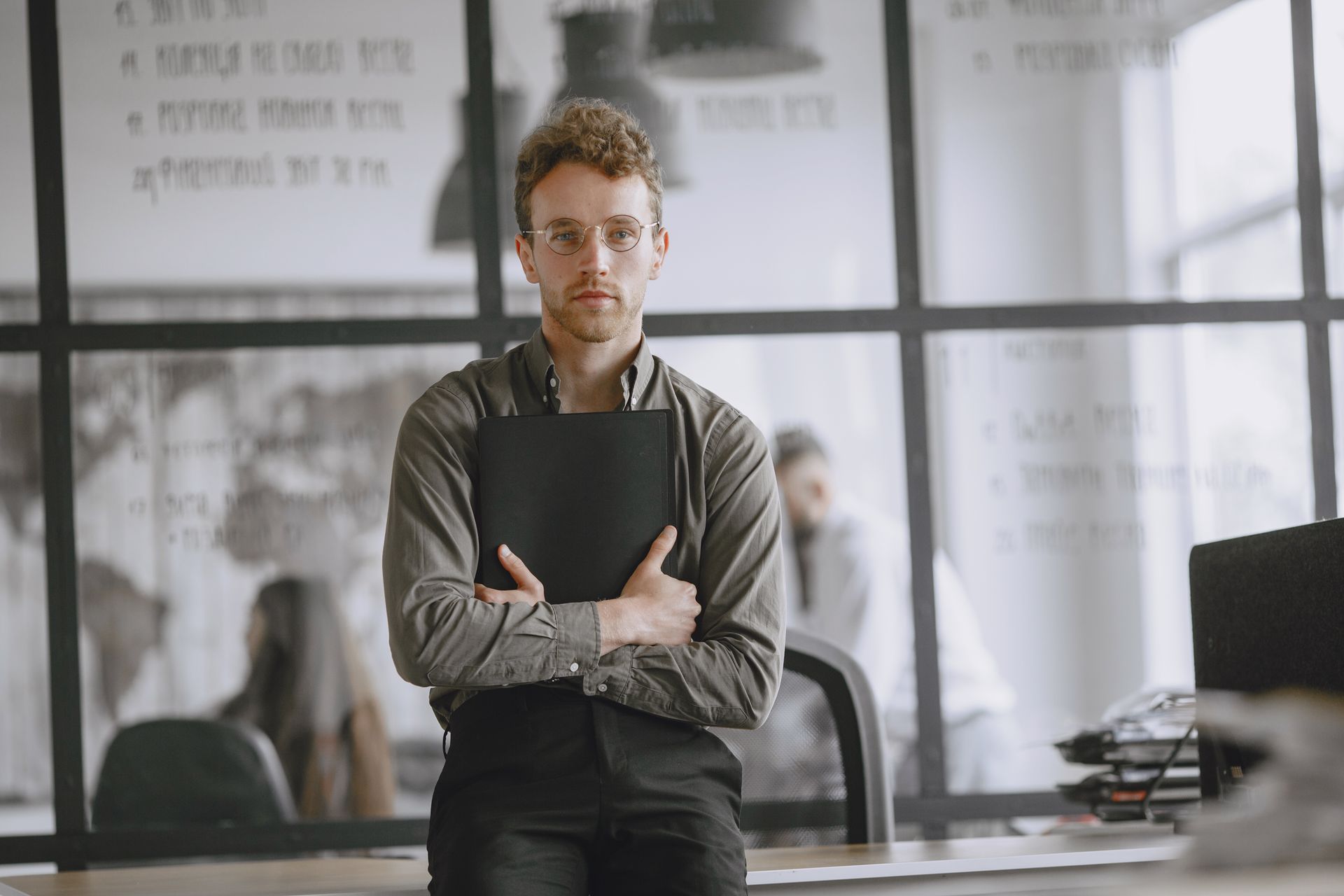 A confident young man standing in a modern office holding a folder, exuding professionalism.