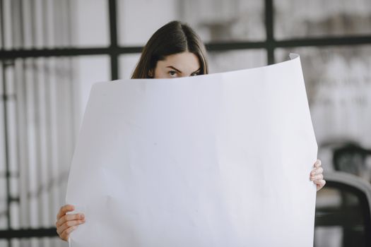A brunette woman partially hides behind a large white paper sheet indoors, looking directly at the camera.
