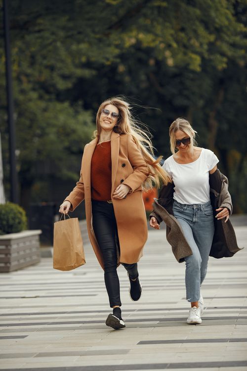 Free Two women running joyfully with shopping bags in a park setting, showcasing fall fashion. Stock Photo