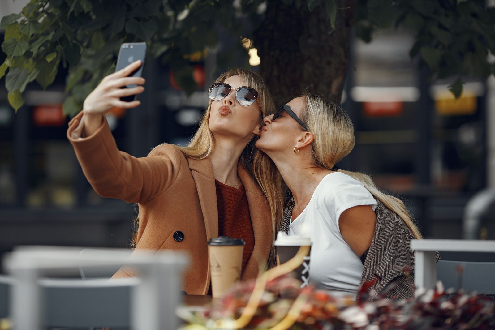 Two friends laughing while taking a fun selfie over coffee outdoors.