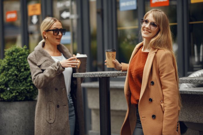 Two women in coats and sunglasses talking over coffee on a city street. Photo by Gustavo Fring on Pexels