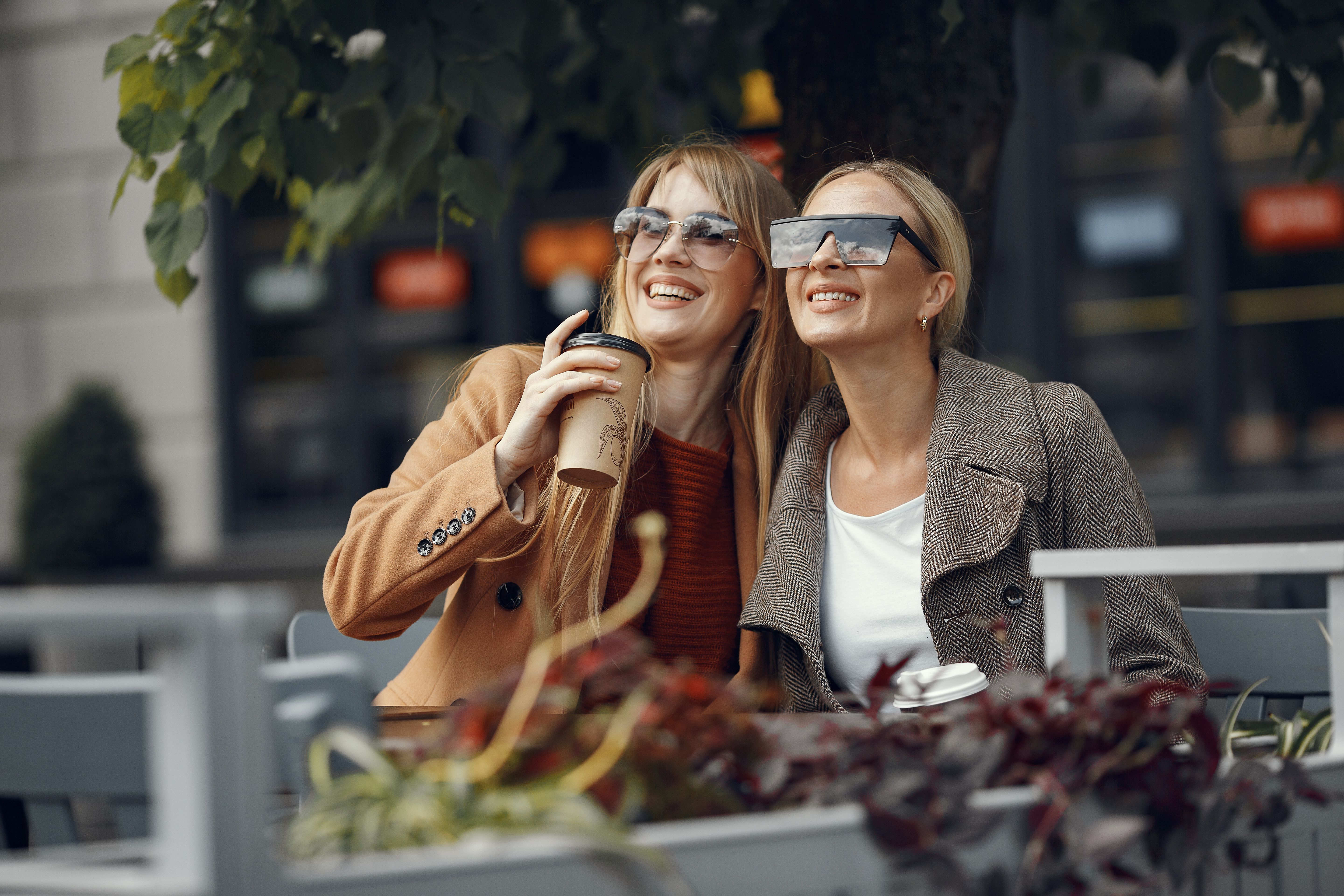 Two women sitting outdoors, smiling and enjoying coffee, wearing sunglasses.
