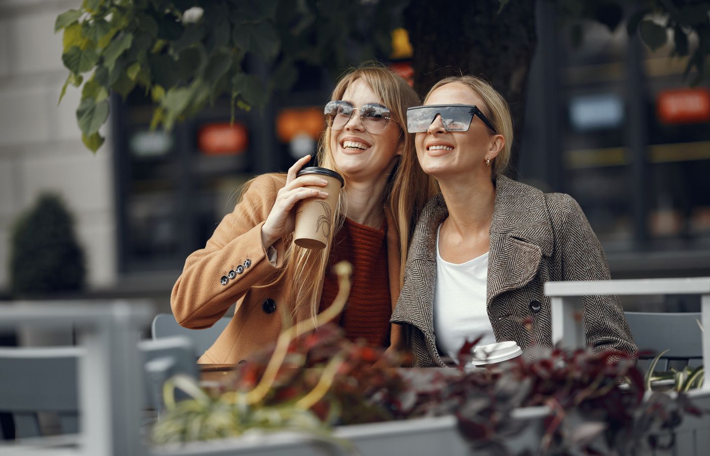 Two women sitting outdoors, smiling and enjoying coffee, wearing sunglasses.