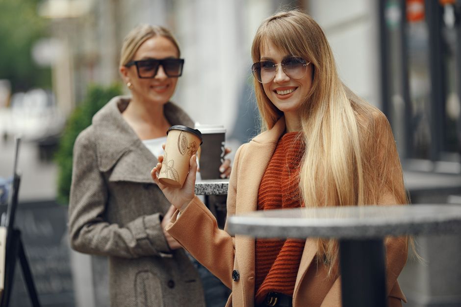 Two women smiling and enjoying coffee on a city street outdoors on a sunny day.