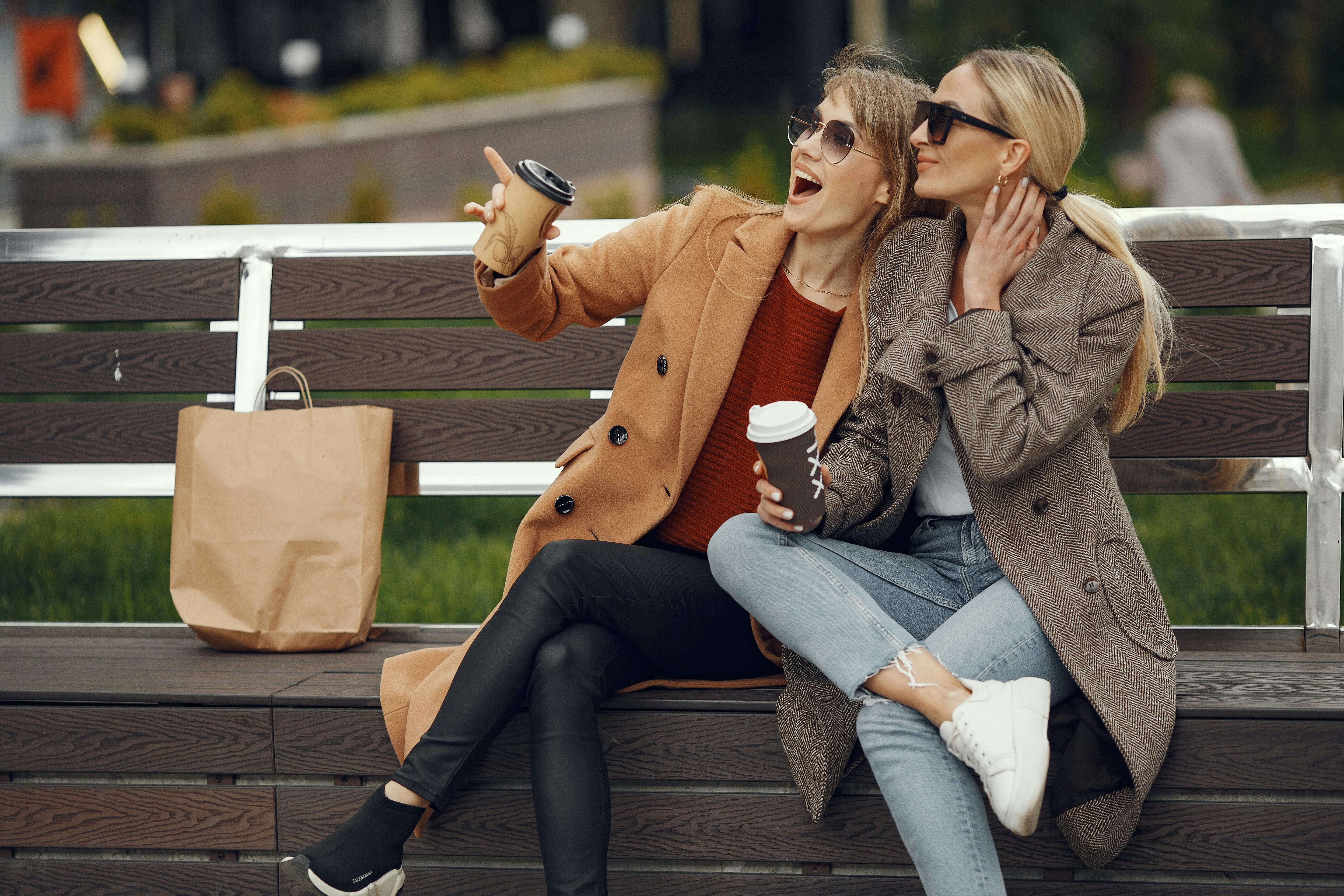 秋日里，两个穿着暖和外套的女人坐在公园长凳上，喝着咖啡开怀大笑