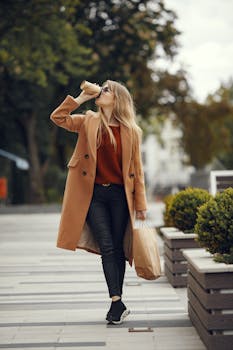Woman in a chic coat enjoys coffee while strolling through a city park, embodying urban lifestyle.