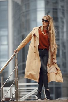 Fashionable woman in a trench coat posing outdoors with urban skyscrapers in the background.