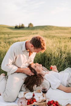 A couple enjoying a romantic picnic in a serene field during sunset, sharing affection and strawberries.