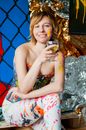 Positive young lady dressed in colorful clothes and paint smears on face sitting on wooden surface with glass of wine and looking at camera during party
